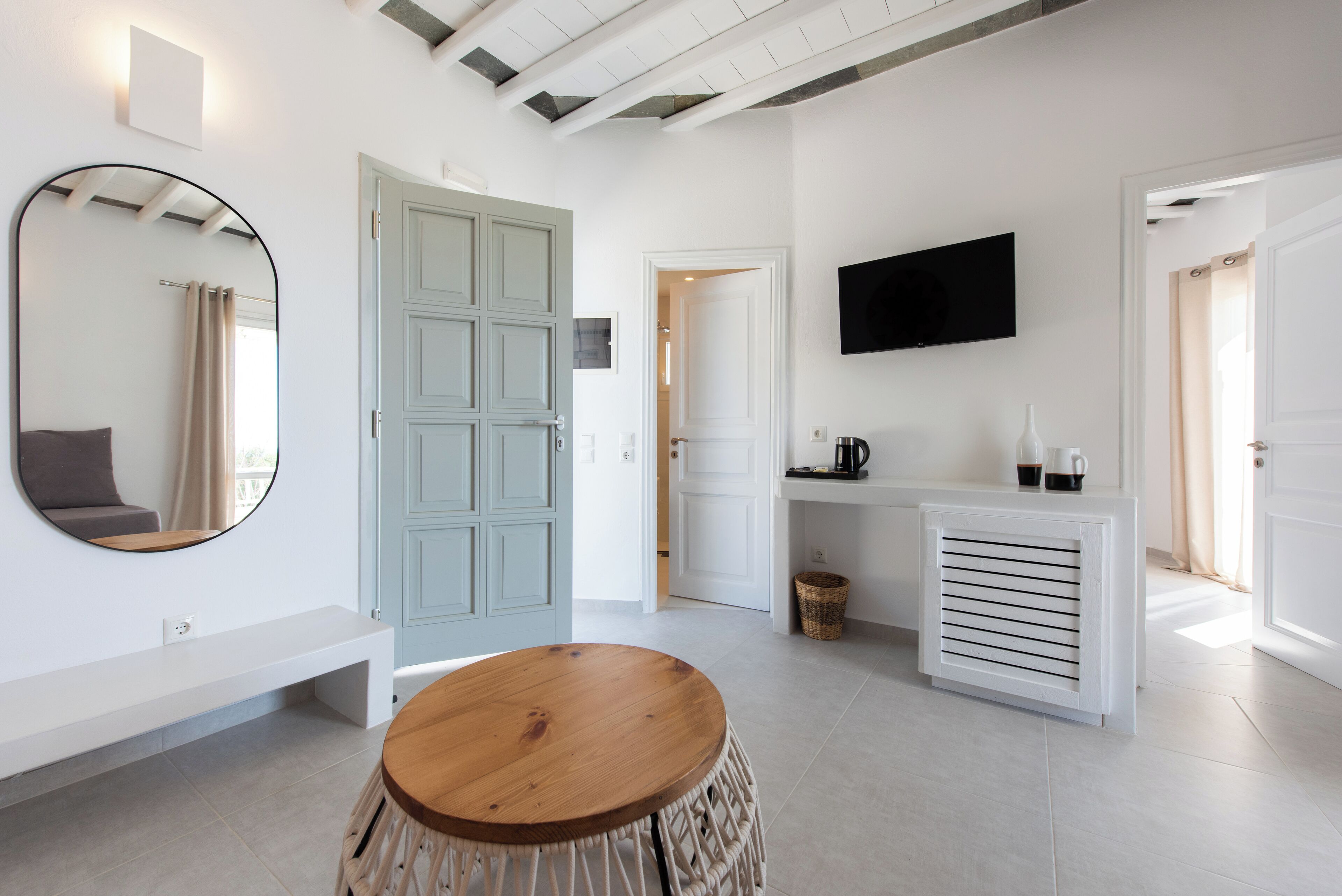 Un intérieur moderne avec une table ronde en bois, un grand miroir, une porte bleu clair, une console blanche avec un téléviseur et un banc blanc.