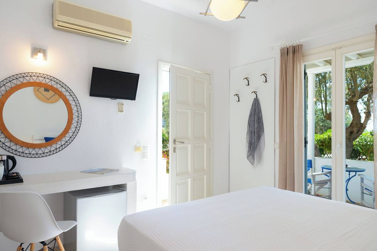 Une chambre d’hôtel comprenant un lit, un bureau avec une chaise, une télévision, un miroir et une porte donnant sur un espace extérieur avec une table et des chaises.