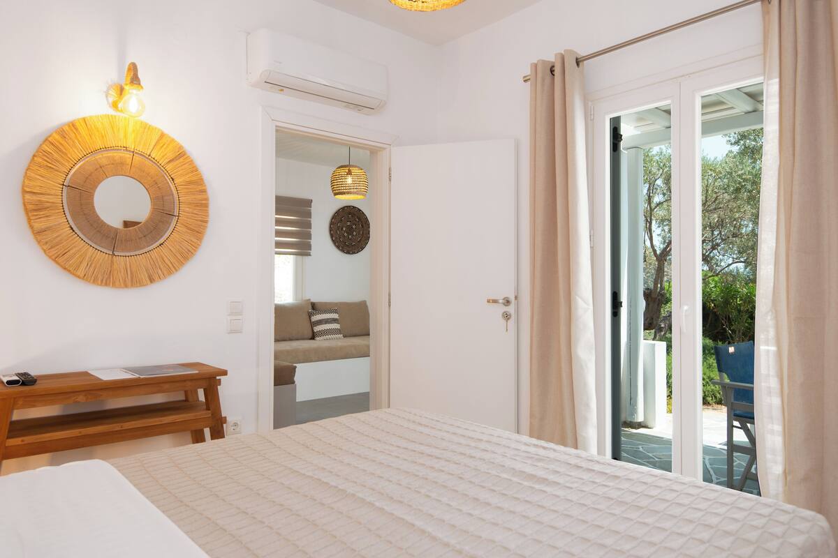 Une chambre à coucher comprenant un lit, une table de chevet en bois, un miroir rond fixé au mur et une porte donnant sur un balcon.