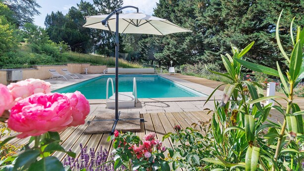 A swimming pool with a wooden deck, surrounded by lush greenery and a large umbrella. - Maison des hôtes – Dordogne: Charming Cottage for 4-5 Guests in Nature (Saint-Crépin-d'Auberoche)