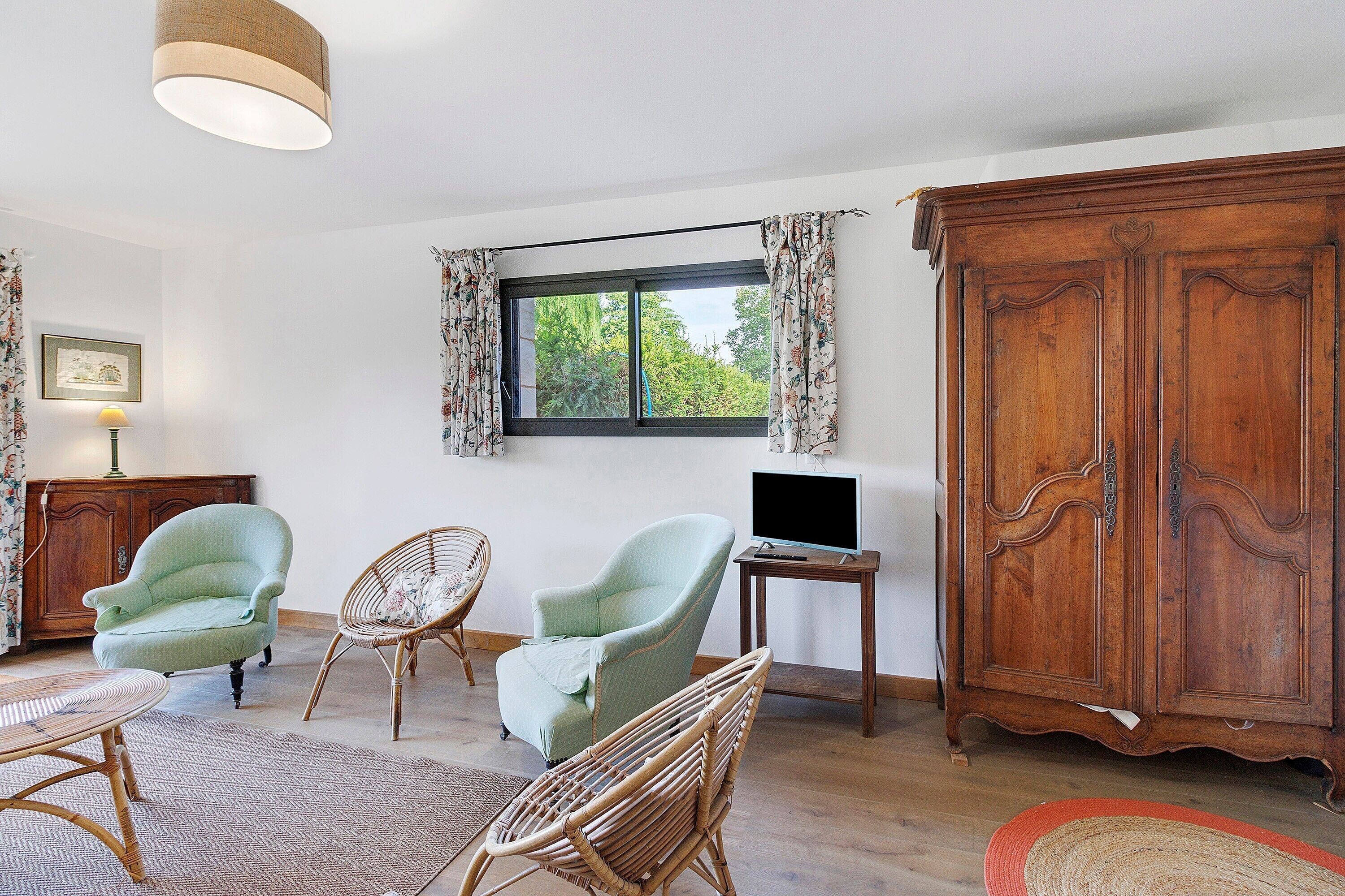Une pièce comprenant une grande armoire en bois, deux fauteuils en osier, une petite table avec une lampe et un téléviseur posé sur une table d’appoint.