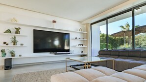 Una sala de estar moderna con un televisor de pantalla plana grande, una mesa de centro de vidrio y un sofá modular. Hay estantes con objetos decorativos y un ventanal con vistas a la vegetación.