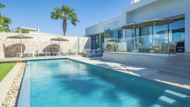 Un área de piscina moderna con agua azul cristalina, paredes de piedra y mobiliario exterior.