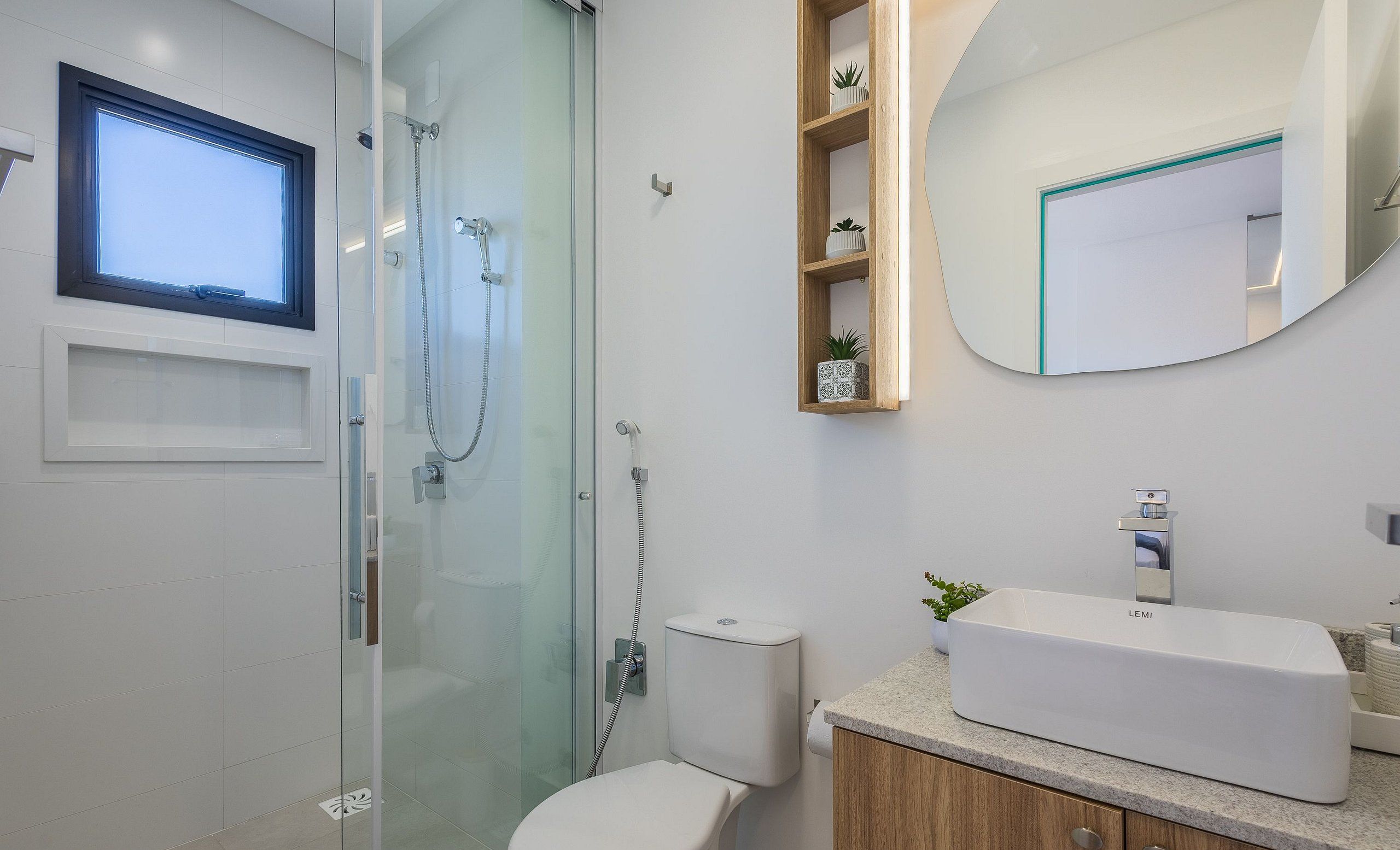 A modern bathroom with a glass-enclosed shower, a toilet, a sink with a LEMI brand soap dispenser, and a mirror with a wooden shelf above it.
