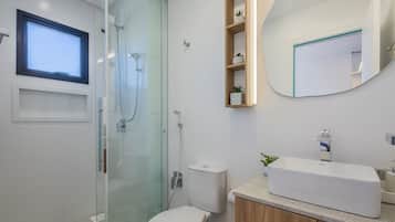 A modern bathroom with a glass-enclosed shower, a toilet, a sink with a LEMI brand soap dispenser, and a mirror with a wooden shelf above it.