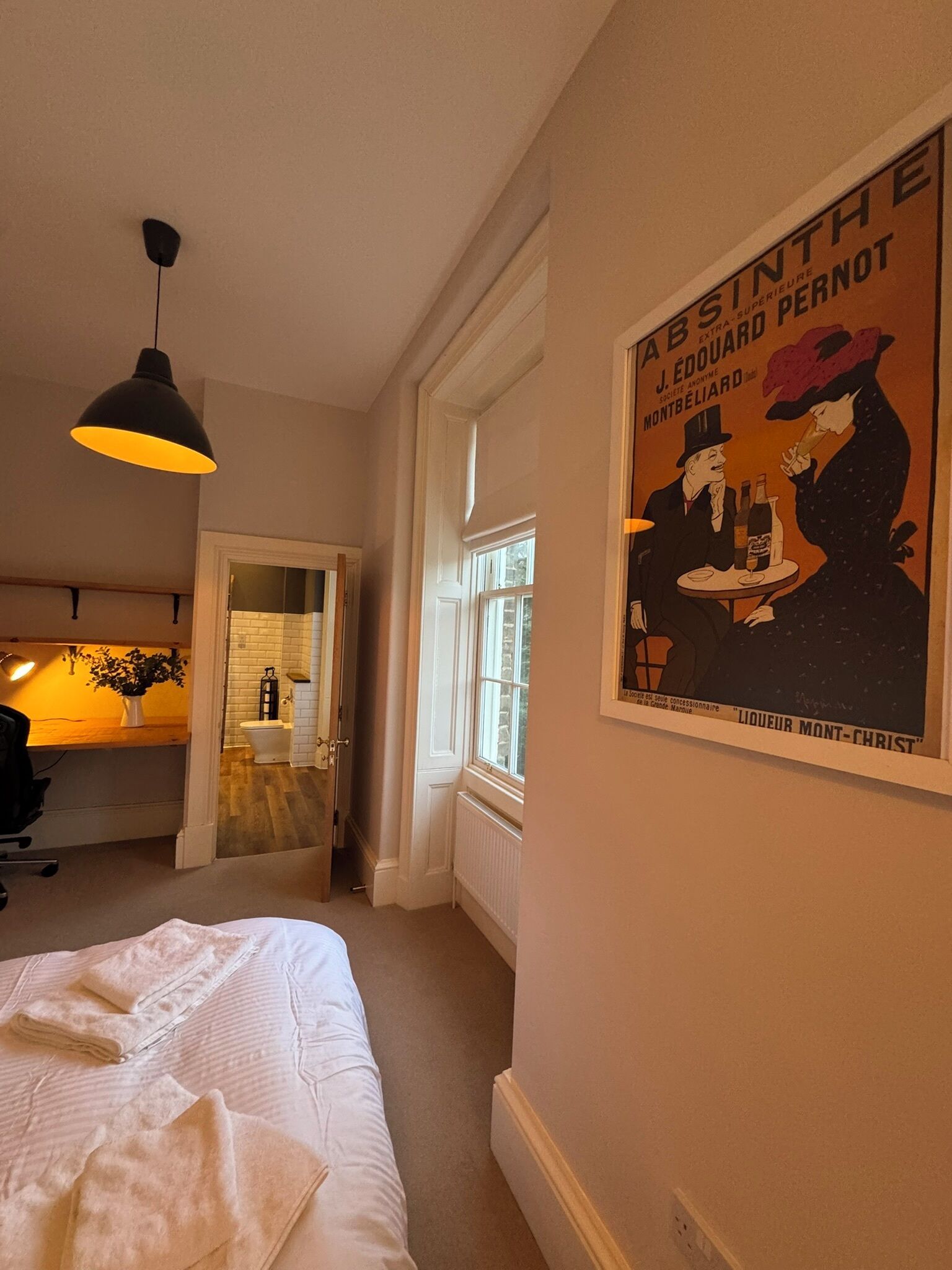 Une chambre d’hôtel avec un lit, un bureau, une chaise, une lampe suspendue, une affiche au mur et une porte donnant sur une salle de bain.