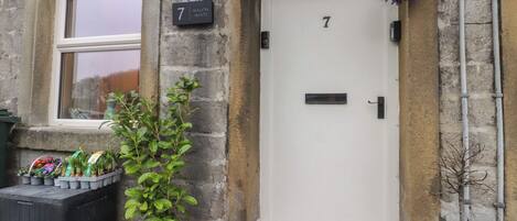 A stone building with a white door marked with the number 7, a small window, and a potted plant in front.