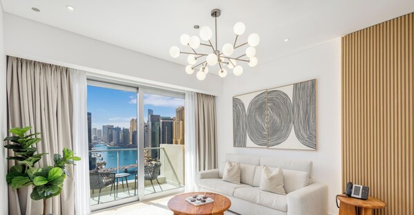A modern living room with a sofa, a coffee table, a plant, and a view of the city through large windows. - LUX The JW Marina Suite 5 (Dubai)