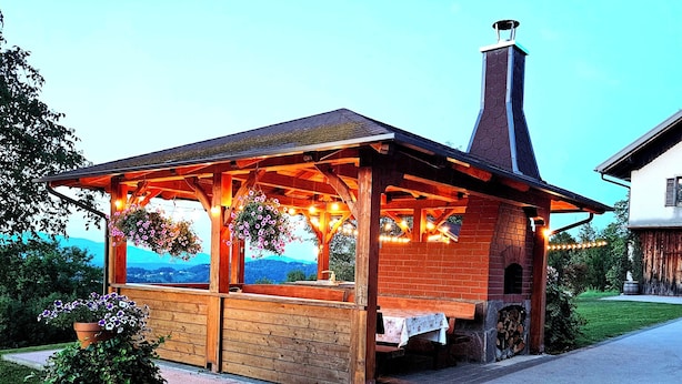 Un kiosque en bois avec une cheminée en briques, une table recouverte d’une nappe blanche et des paniers de fleurs suspendus.