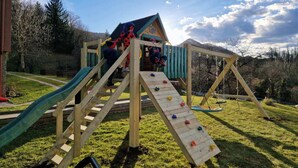 Une structure de jeux en bois, dotée d’un toboggan et d’un mur d’escalade, installée dans un espace verdoyant, agrémenté d’arbres et offrant une vue sur un ciel dégagé.
