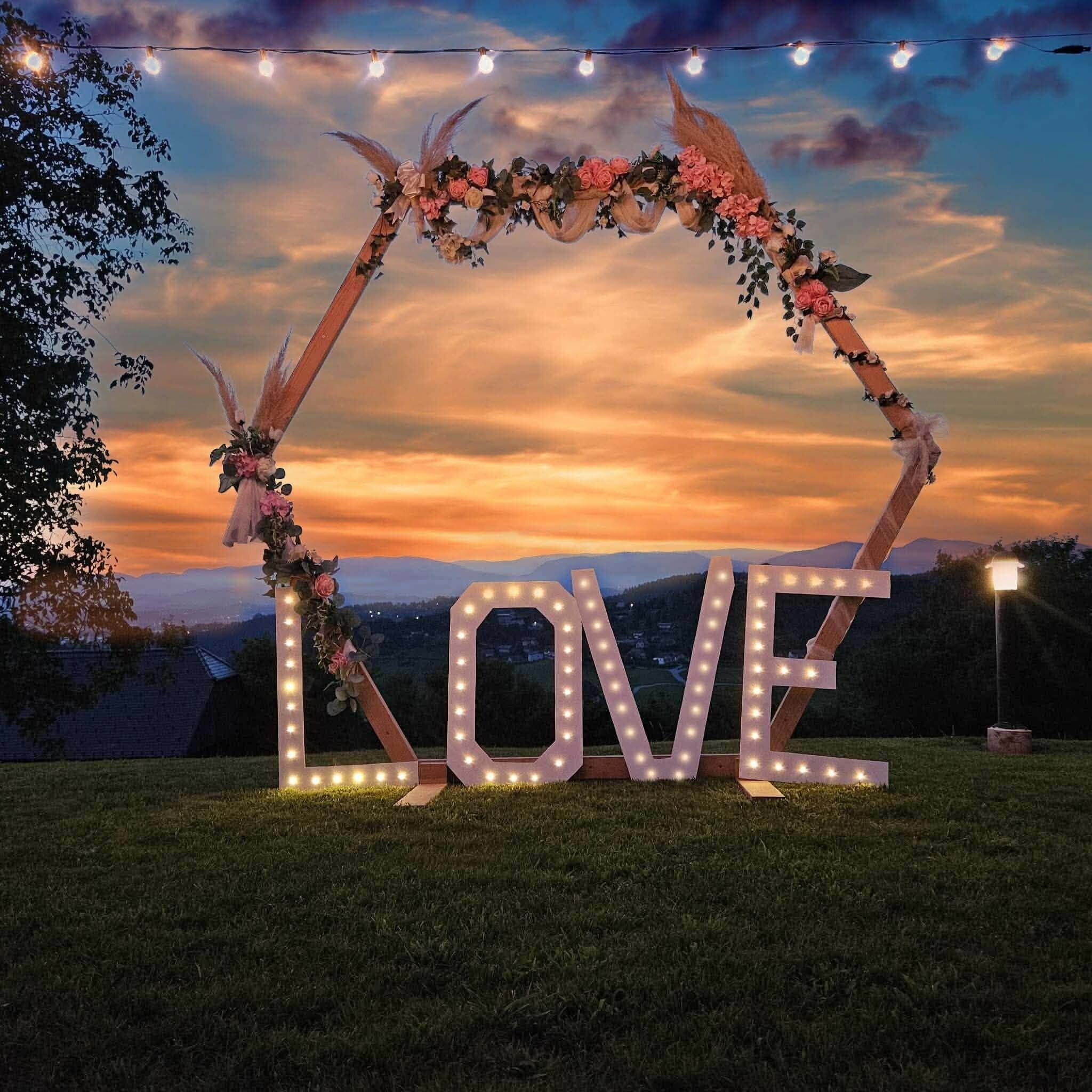 An outdoor arch with the word "LOVE" illuminated, surrounded by string lights and floral decorations against a sunset backdrop.