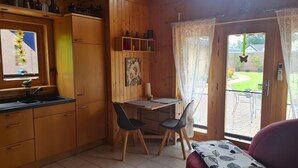 Una cocina compacta con armarios de madera, un comedor con dos sillas, una mesita y un ventanal con vistas a un jardín.