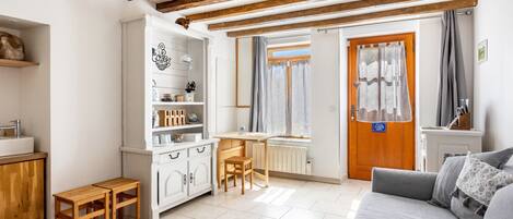 A room with a wooden ceiling, a white cabinet, a wooden table with a chair, a radiator, and a grey sofa.