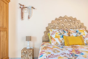 Een slaapkamer met een bloemenbedekking, een houten hoofdbord, een nachtkastje met een lamp en een macramé decoratie aan de muur.