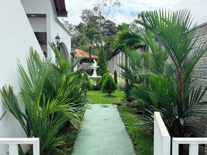 A narrow pathway flanked by lush greenery and a white fence, leading to a building with a tiled roof.