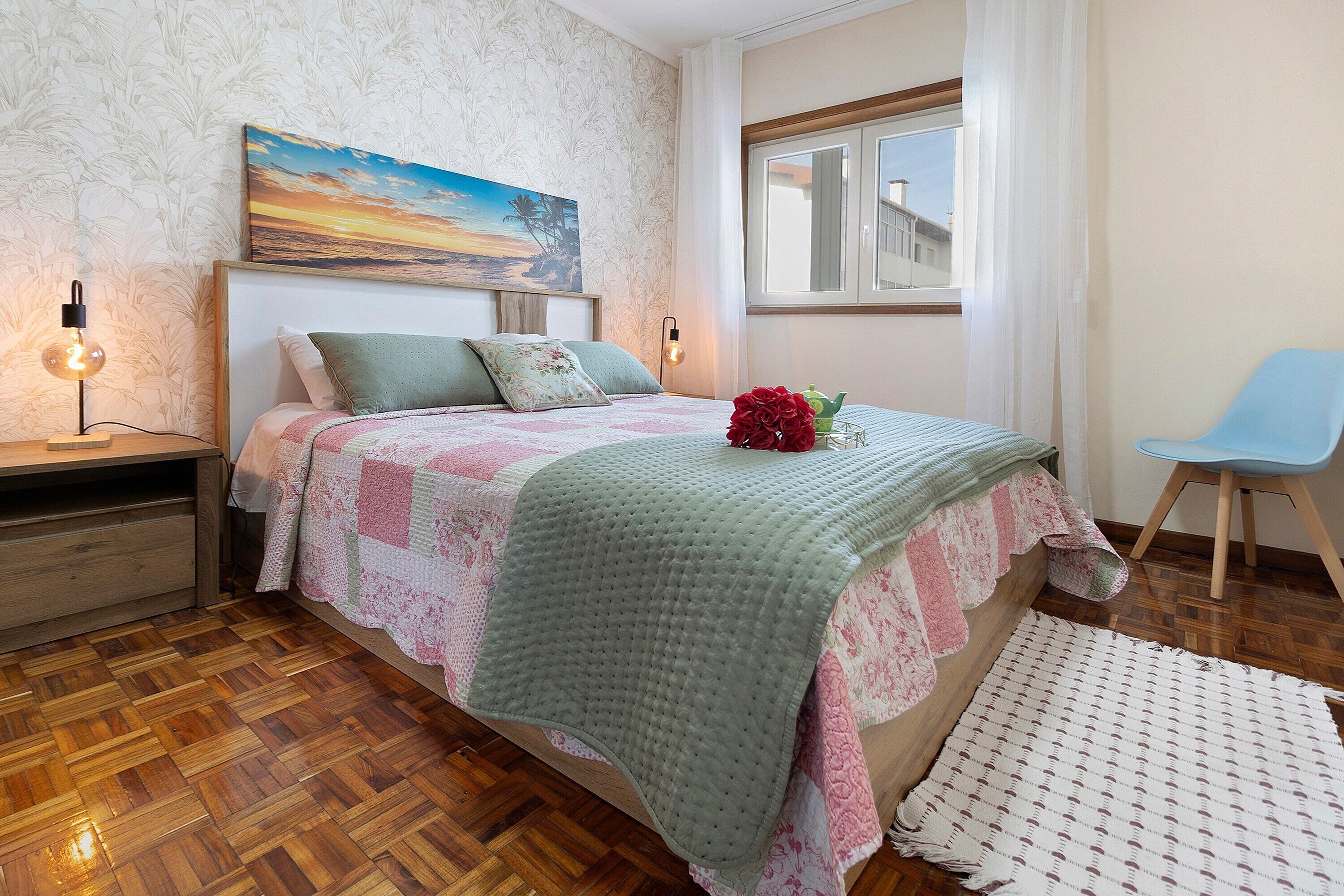 A bedroom with a wooden floor, a bed with a quilt, a nightstand, a chair, and a window with a view.