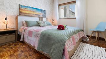 A bedroom with a wooden floor, a bed with a quilt, a nightstand, a chair, and a window with a view.