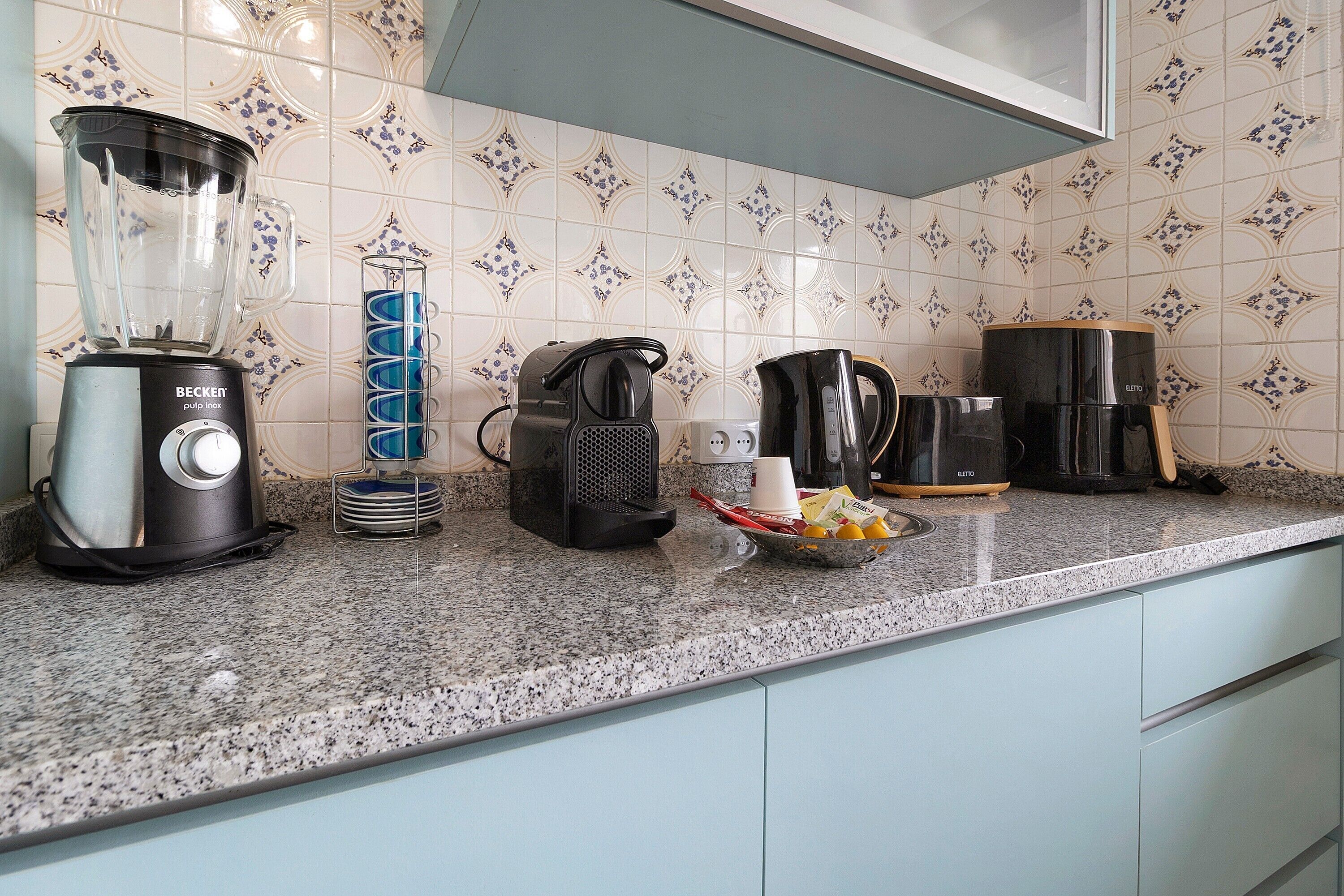 A kitchen with a blender, coffee maker, kettle, and a bowl with cups and condiments.