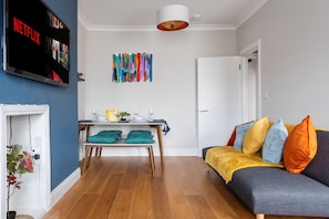 A living room with a TV displaying Netflix, a dining table with a plant, and a sofa with colorful cushions. - Vibrant & Stylish|2BR|Shepherd’s Bush|Fast WiFi|ST (Greater London)