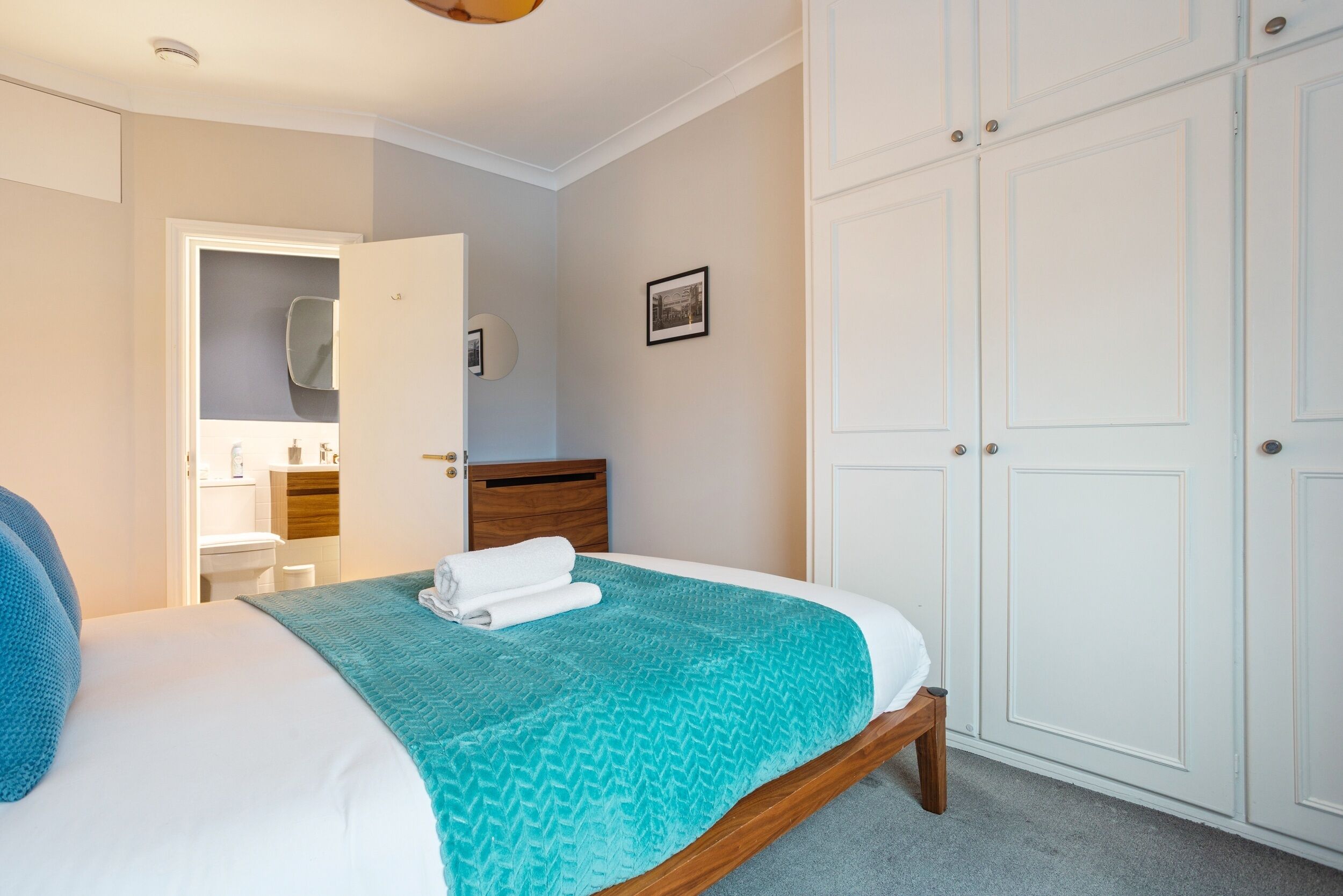 Une chambre à coucher avec un lit, une tête de lit en bois, une couvre-lit bleue et blanche, une porte blanche donnant accès à une salle de bain et une armoire blanche.