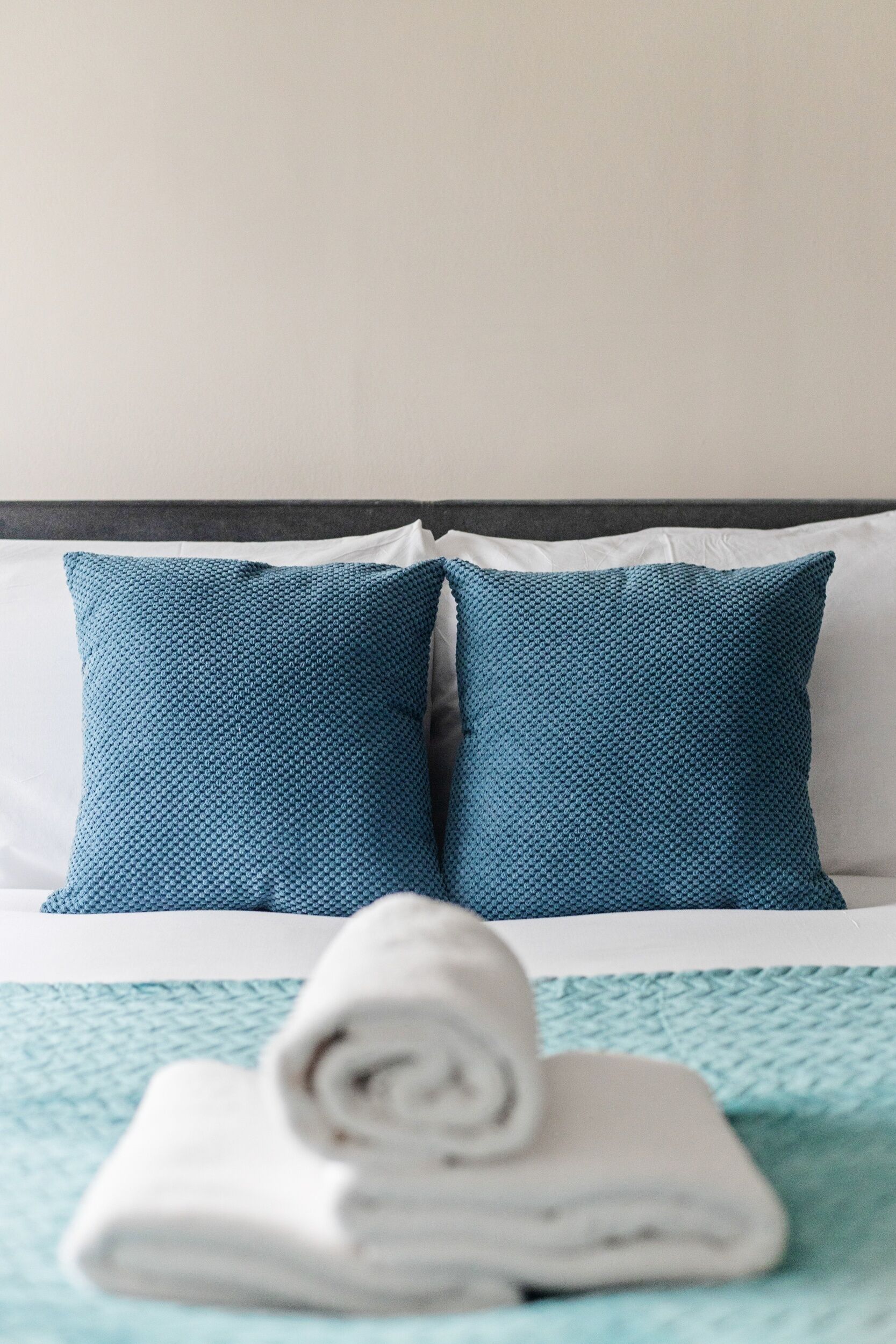 Ein ordentlich gemachtes Bett mit zwei blauen Kissen und einem am Fuß gerollten weißen Handtuch.