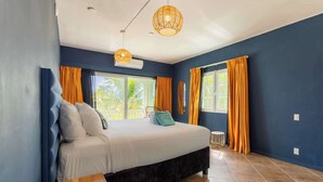 A bedroom with a bed, a bedside table, a hanging light fixture, and a window with curtains. - Modern Apt w/ Sea View & Pool | Jan Thiel (Willemstad)
