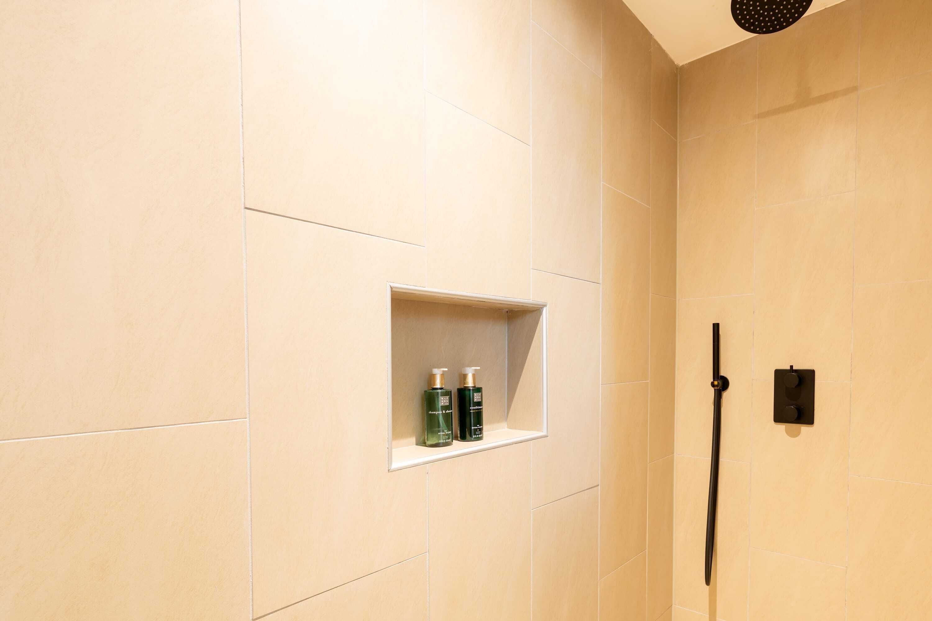 Baño con azulejos color crema, cabezal de ducha y estante con dos botellas.