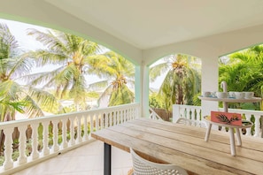 A wooden table with a white chair, a small white table with cups and a card, and a balcony with white railings and palm trees. - Modern Apt w/ Plunge Pool | Jan Thiel (Willemstad)