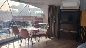 Een kamer met een groot raam, een ronde tafel met stoelen, een televisie en een plafondventilator.