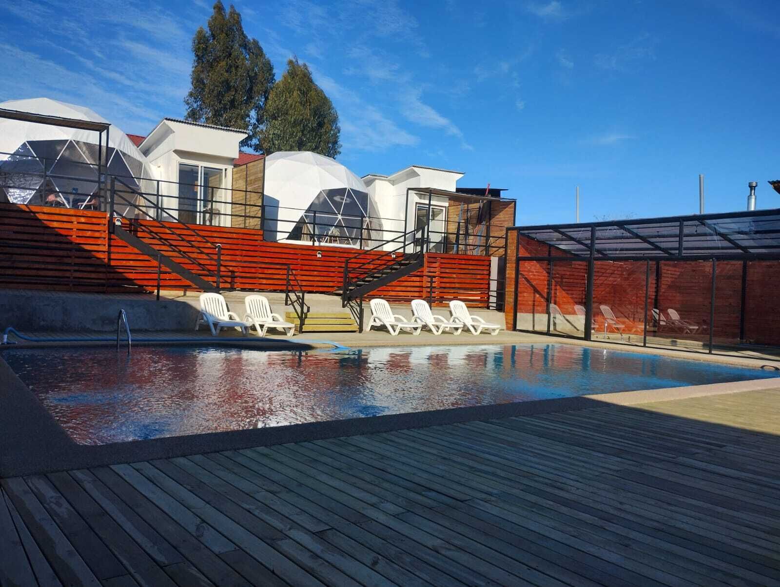 Una piscina con sillas de descanso, una zona cubierta y un edificio de diseño moderno con detalles en rojo.