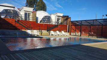 Una piscina con sillas de descanso, una zona cubierta y un edificio de diseño moderno con detalles en rojo.