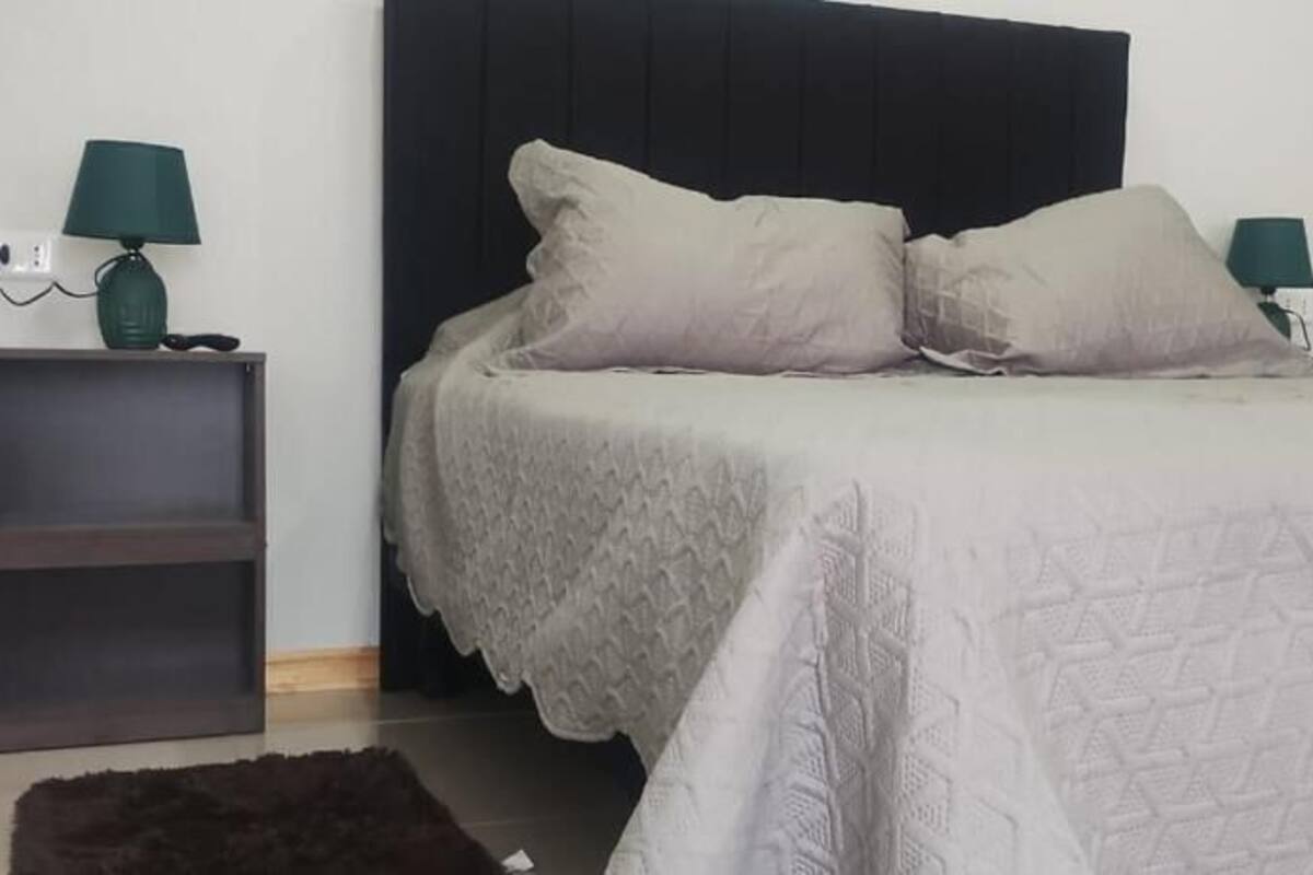 Una cama bien hecha con una colcha blanca y dos almohadas, flanqueada por dos mesitas de noche con lámparas, en una habitación sencilla con cabecera oscura.