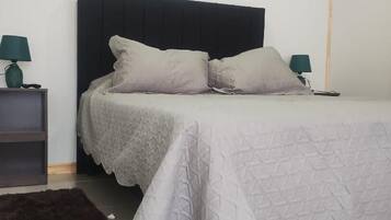 Un lit bien fait, recouvert d’une couvre-lit blanche et agrémenté de deux oreillers, flanqué de deux tables de chevet avec des lampes, dans une chambre simple avec une tête de lit sombre.