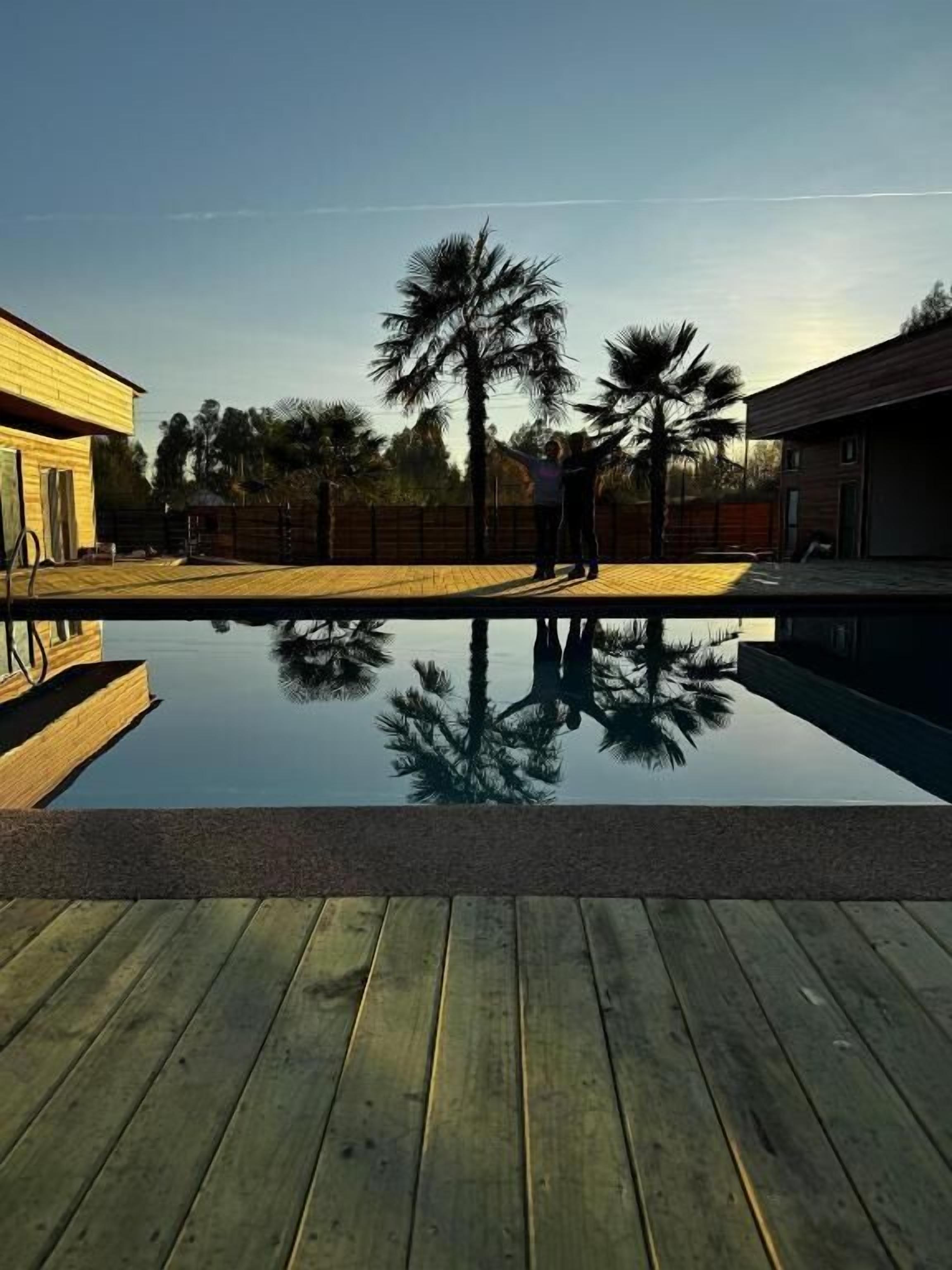 Eine Holzterrasse mit Swimmingpool, der Palmen und ein Gebäude im Hintergrund spiegelt.
