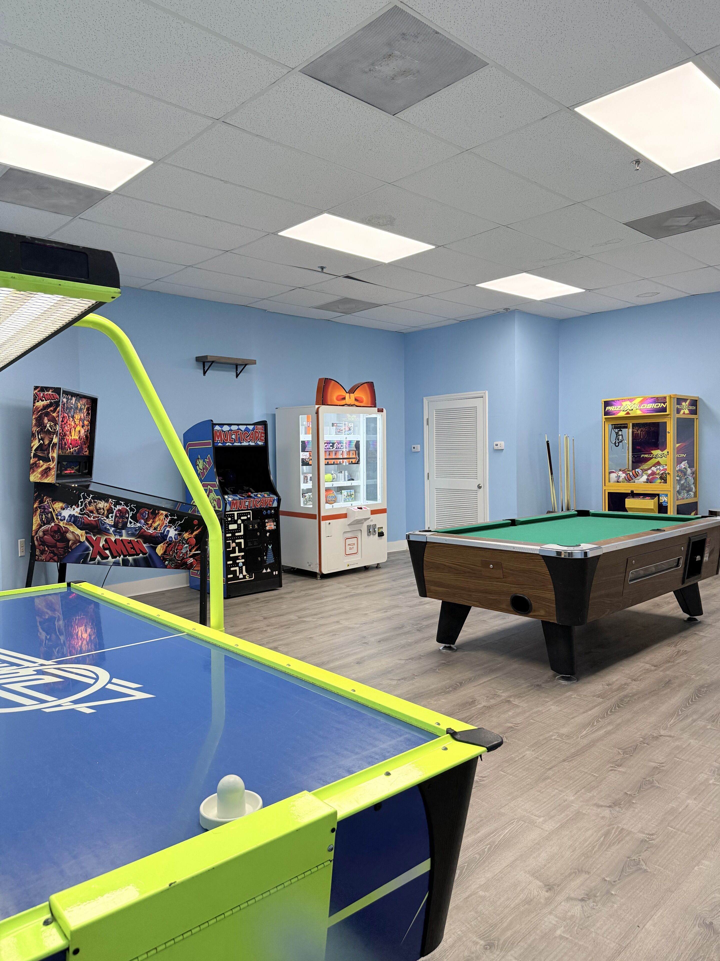A pool table, arcade machines, and a vending machine in a room with blue walls and a wooden floor.