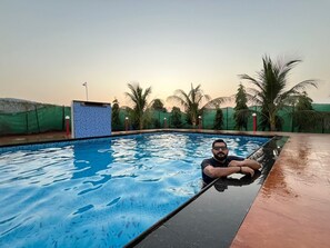 A swimming pool with clear blue water, surrounded by palm trees and a green fence. - Shravani Farms  (Tala)