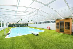 Un espace piscine doté d’un toit en verre, d’un revêtement d’herbe synthétique et d’une petite structure en bois.