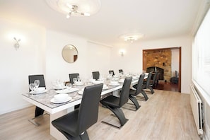 Une salle à manger avec une longue table dressée pour un repas, des chaises noires, un grand miroir rond et une cheminée en pierre.