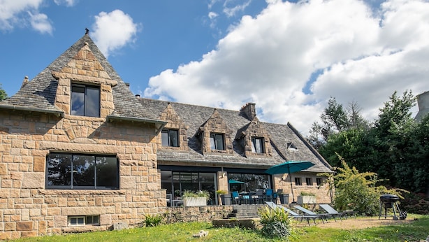Ein Steinhaus mit einem steilen Dach, großen Fenstern und einer überdachten Terrasse mit Gartenmöbeln.