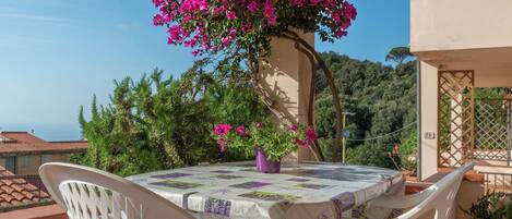 A round table with a patterned tablecloth, two white chairs, and a potted plant with pink flowers.