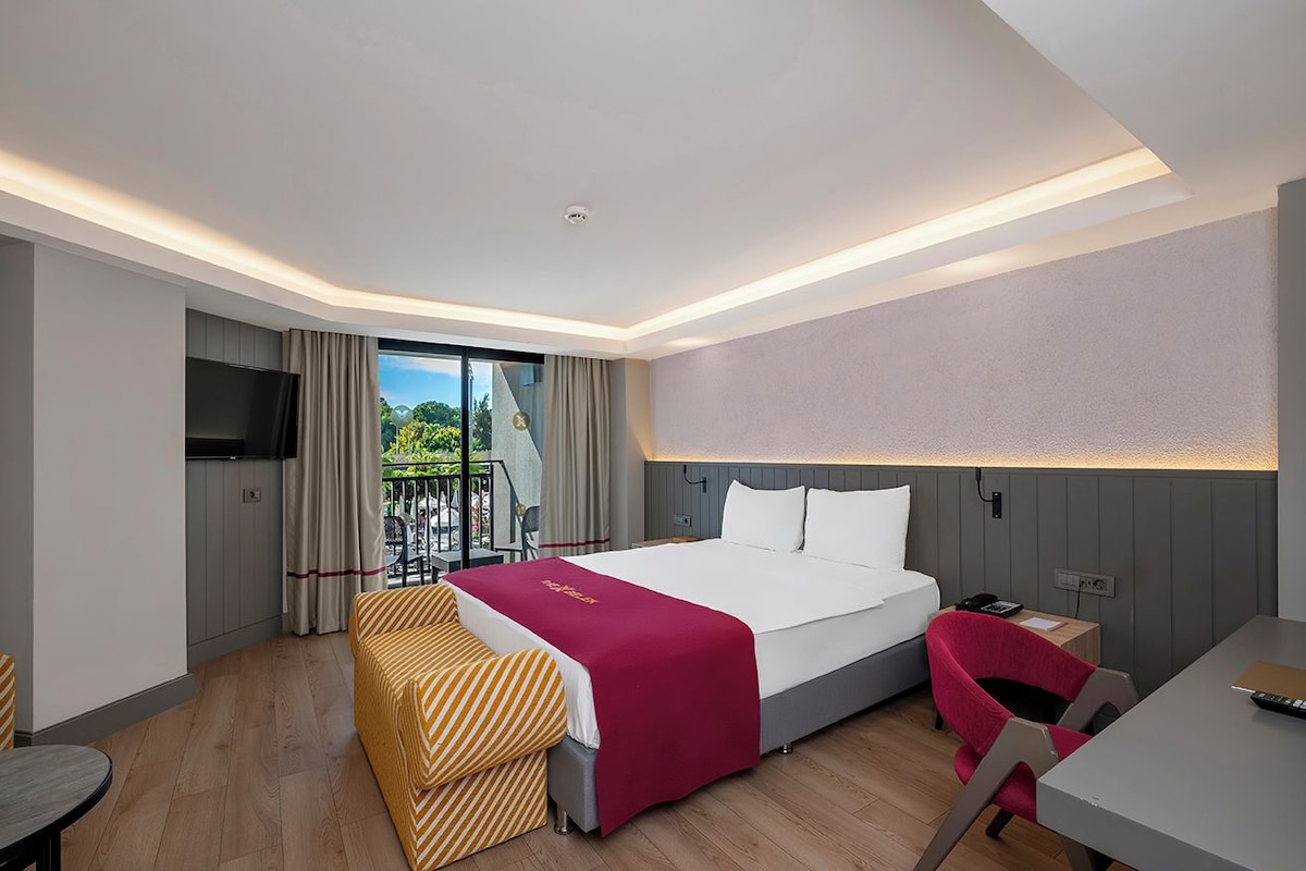 A modern hotel room with a large bed, a desk, a chair, a TV, and a balcony with a view of greenery.