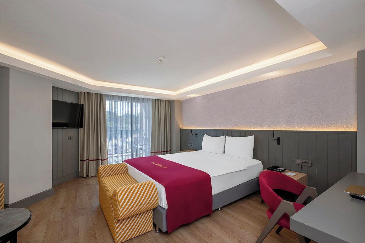A modern hotel room with a large bed, a desk, a chair, a TV, and a window with curtains.