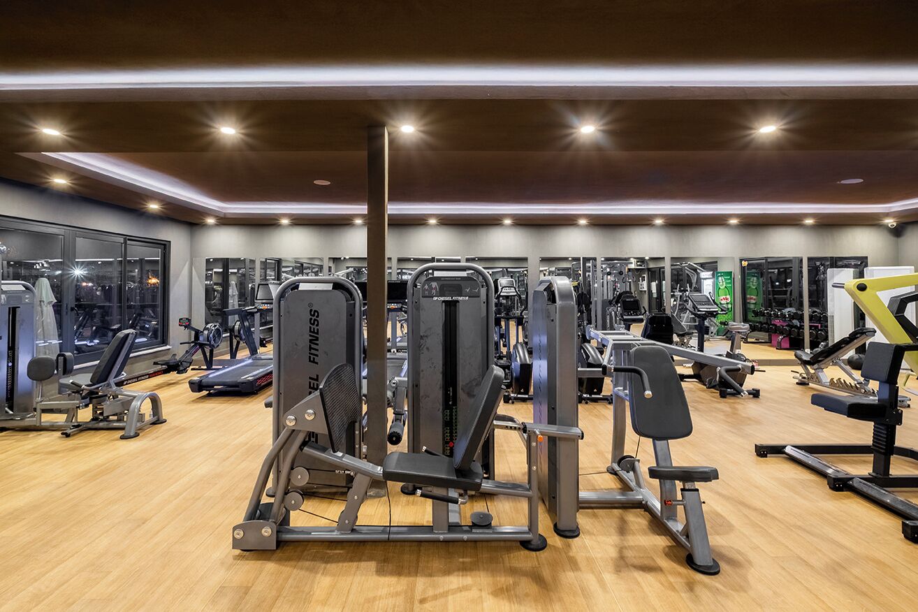 Un gimnasio con diversas máquinas de ejercicio, amplias ventanas y suelo de madera.