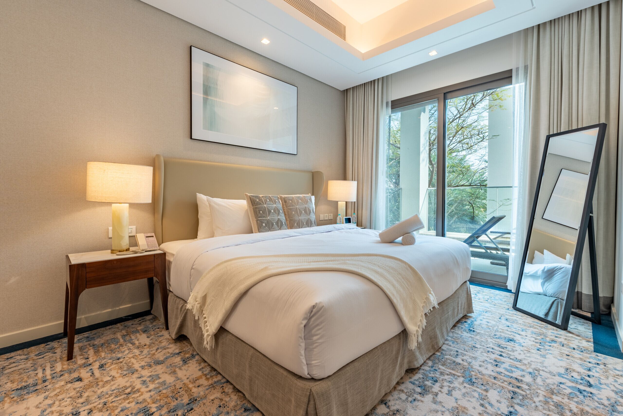 A hotel room with a large bed, two bedside lamps, a bedside phone, a large window with a view of the outdoors, and a ceiling light.