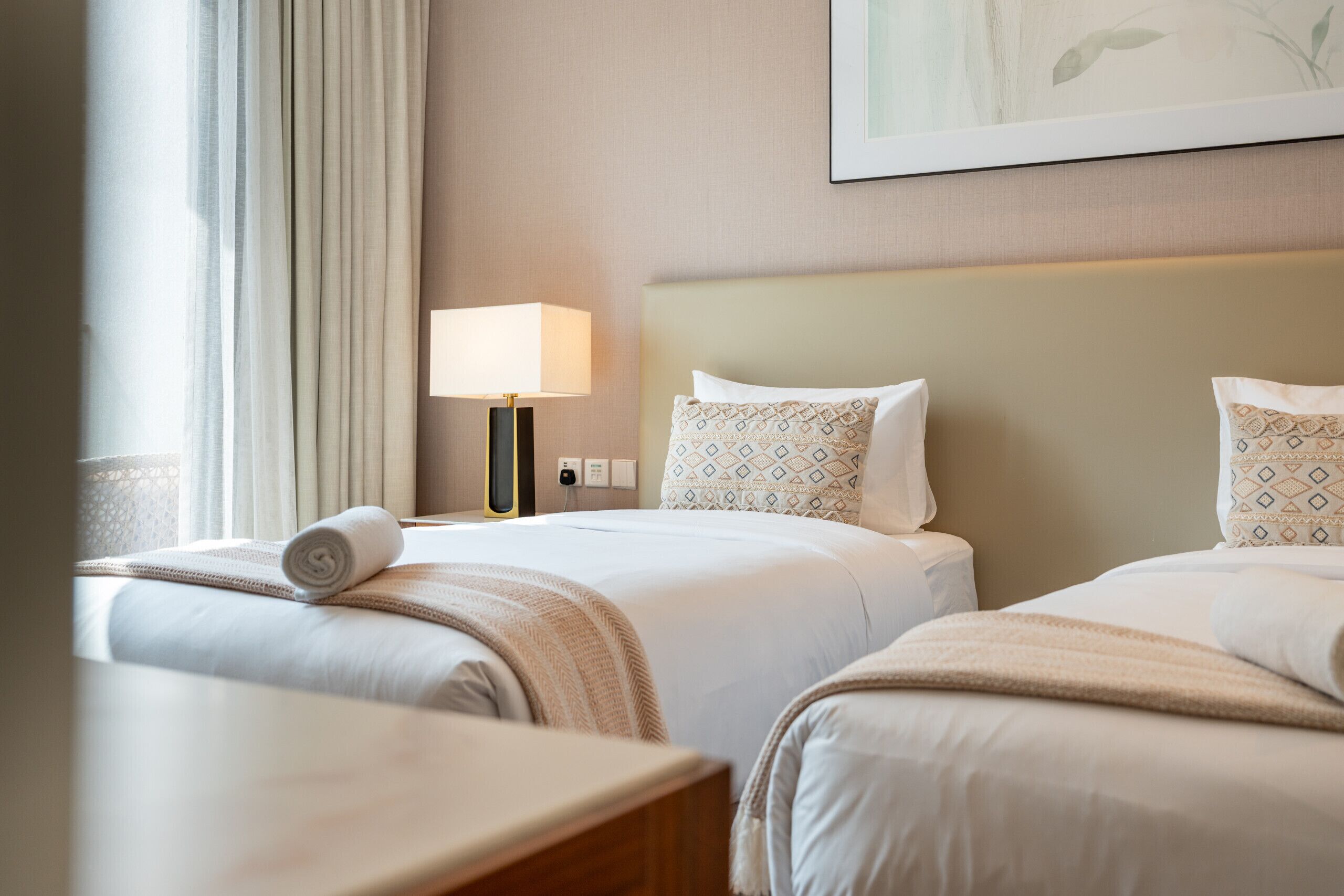 A hotel room with two beds, a bedside table, a lamp, and a painting on the wall.