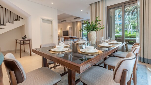 A modern dining area with a long table set for six, a staircase, and a view of the outdoors. - Peaceful Villa Bedroom + Assistant Room, Cliffside View (Fujairah)