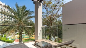 A balcony with wicker furniture, a glass table, and a view of a landscaped area with palm trees and a playground. - Peaceful Villa Bedroom + Assistant Room, Cliffside View (Fujairah)