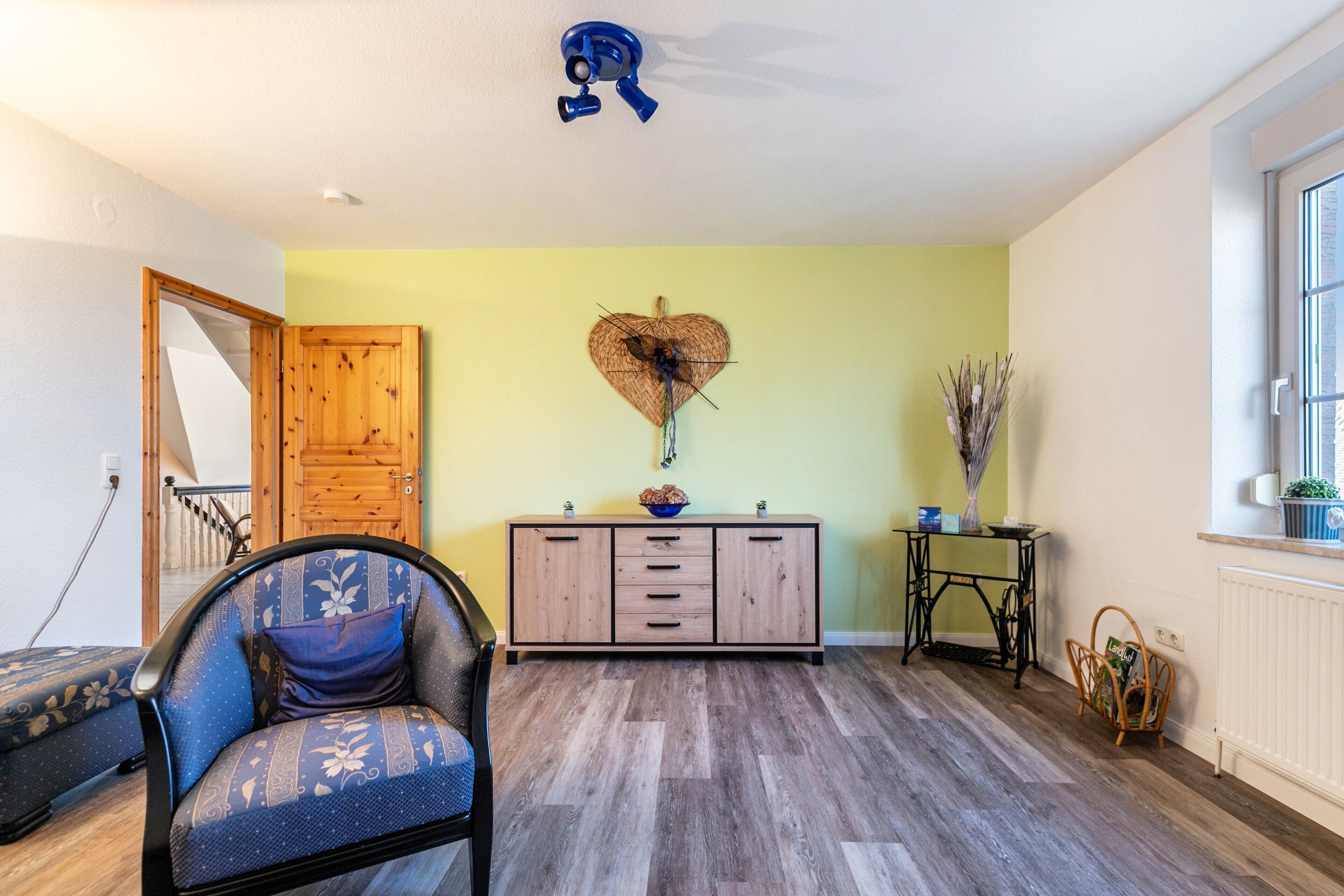 A room with a wooden cabinet, a blue upholstered chair, a heart-shaped wall decoration, and a window.