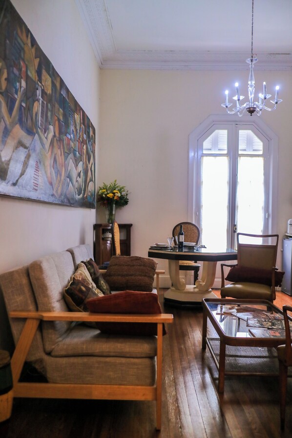 A living room with a wooden sofa, a dining table with chairs, a chandelier, and a large abstract painting on the wall.