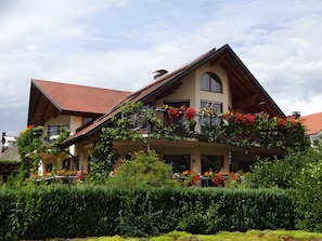 A two-story house with a sloped roof, a balcony with red flowers, and a well-maintained garden.